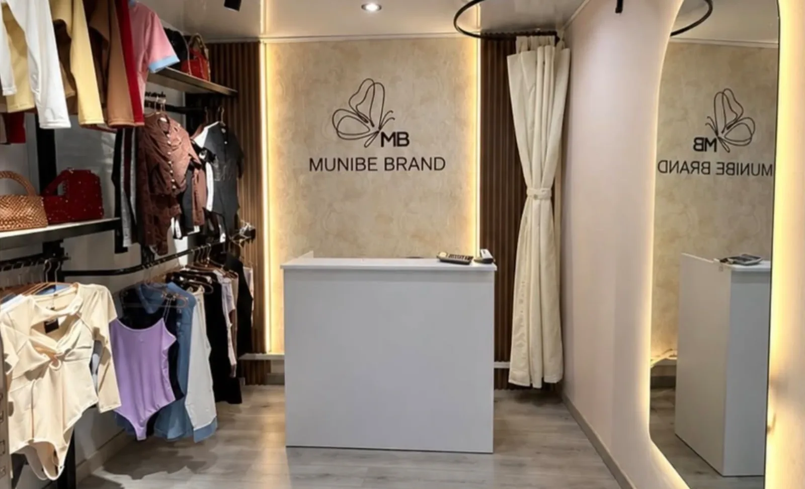 Interior de la tienda Munibe Brand en Medellín: percheros con prendas, el mostrador con el logo en la pared, el probador con cortina y un espejo en arco iluminado.